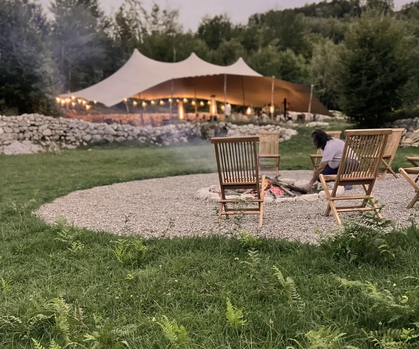 The fire circle on the meadow at ašuma retreat at dusk, with the lit pavilion behind the stone wall