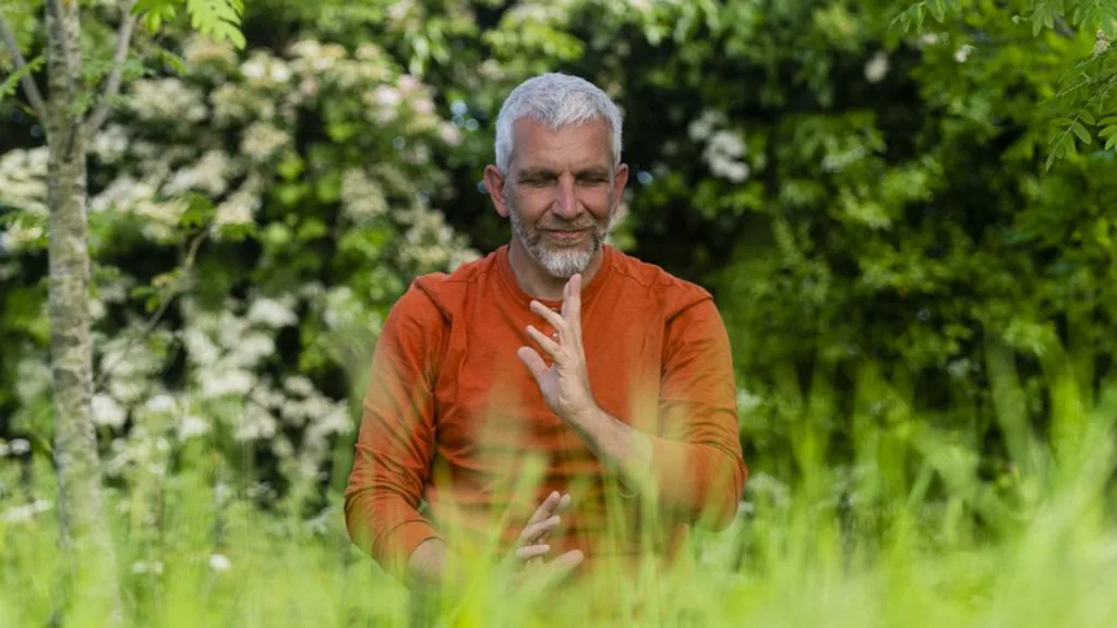 John Millar in an orange top among tall grass and blossom, hands in front of his body